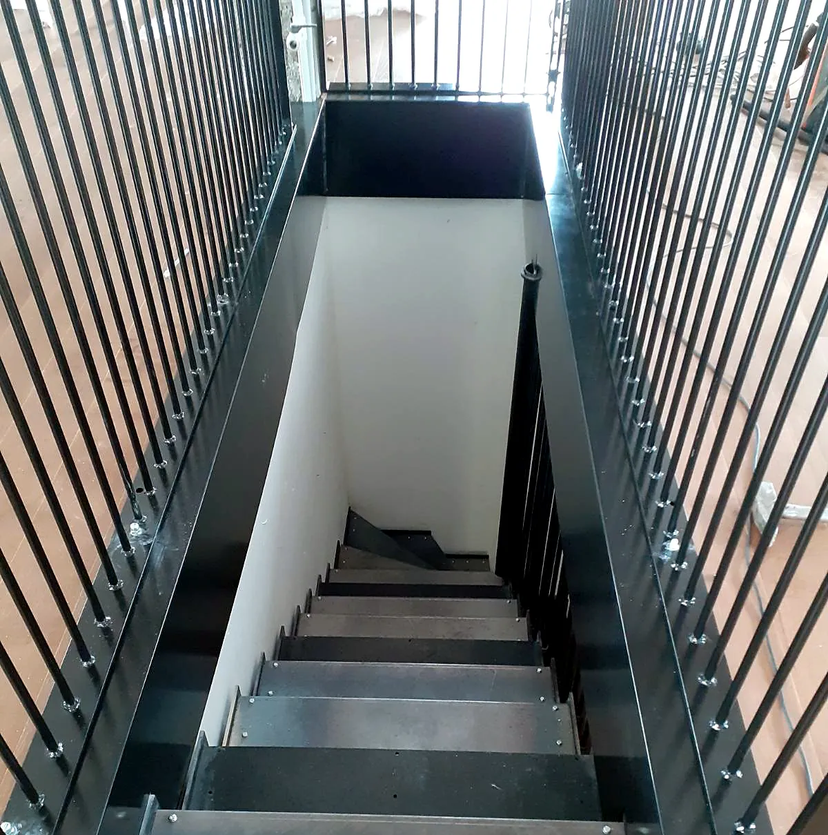 Stahltreppe in einer Deckenöffnung, von oben zwischen zwei senkrechten Stabgeländern